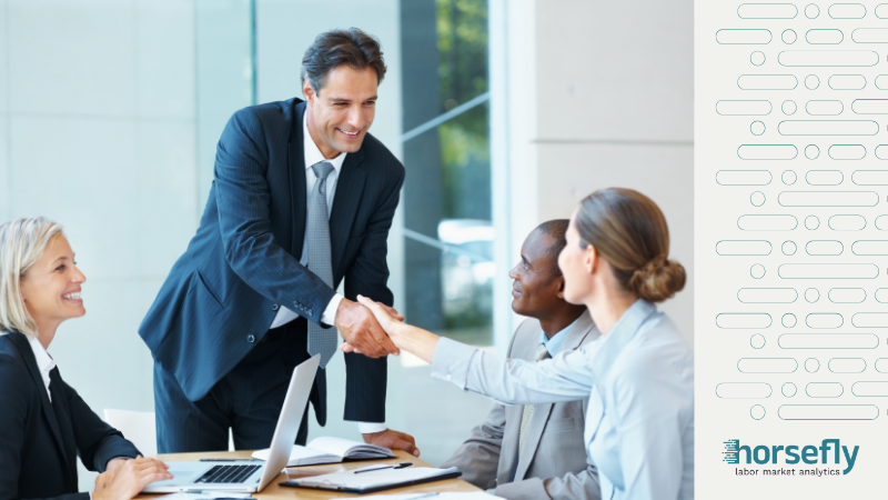 Image shows a group of business people around a table, with a business man standing and shaking hands with a business woman at the table - for the blog - Shifting the Focus: Skills Over Experience in Recruitment